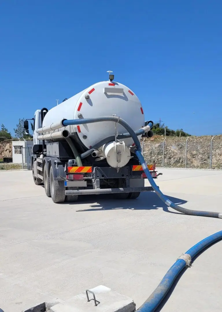 A large tanker truck with hoses connected is parked on a concrete surface under a clear blue sky.