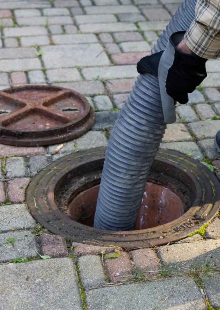 A person inserts a large hose into an open manhole on a brick-paved surface.