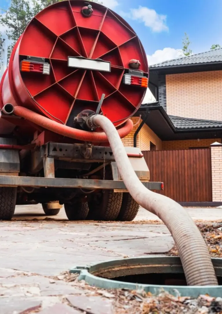 A red tanker truck is parked near a manhole, and a hose extends from the truck into the manhole, suggesting sewage or waste removal at a residential property.