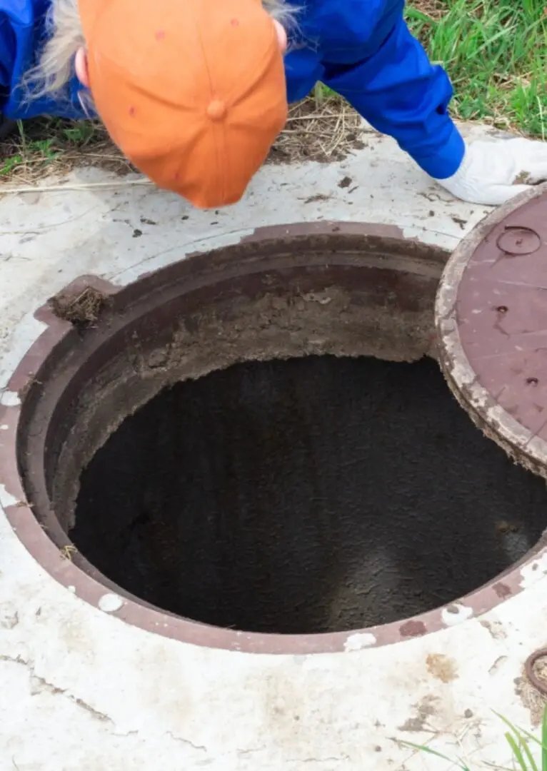 A person wearing a blue outfit and orange cap peers into an open manhole.