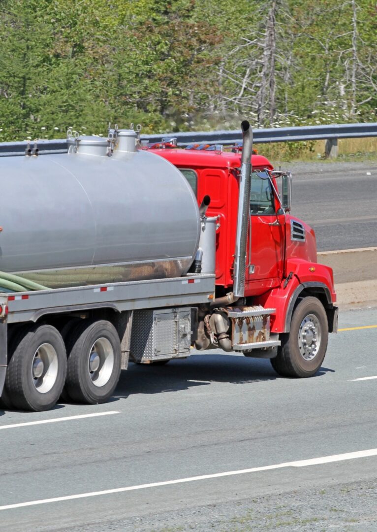 A red tanker truck drives on a highway with a metal tank attached behind the cab. Trees and a barrier can be seen in the background.